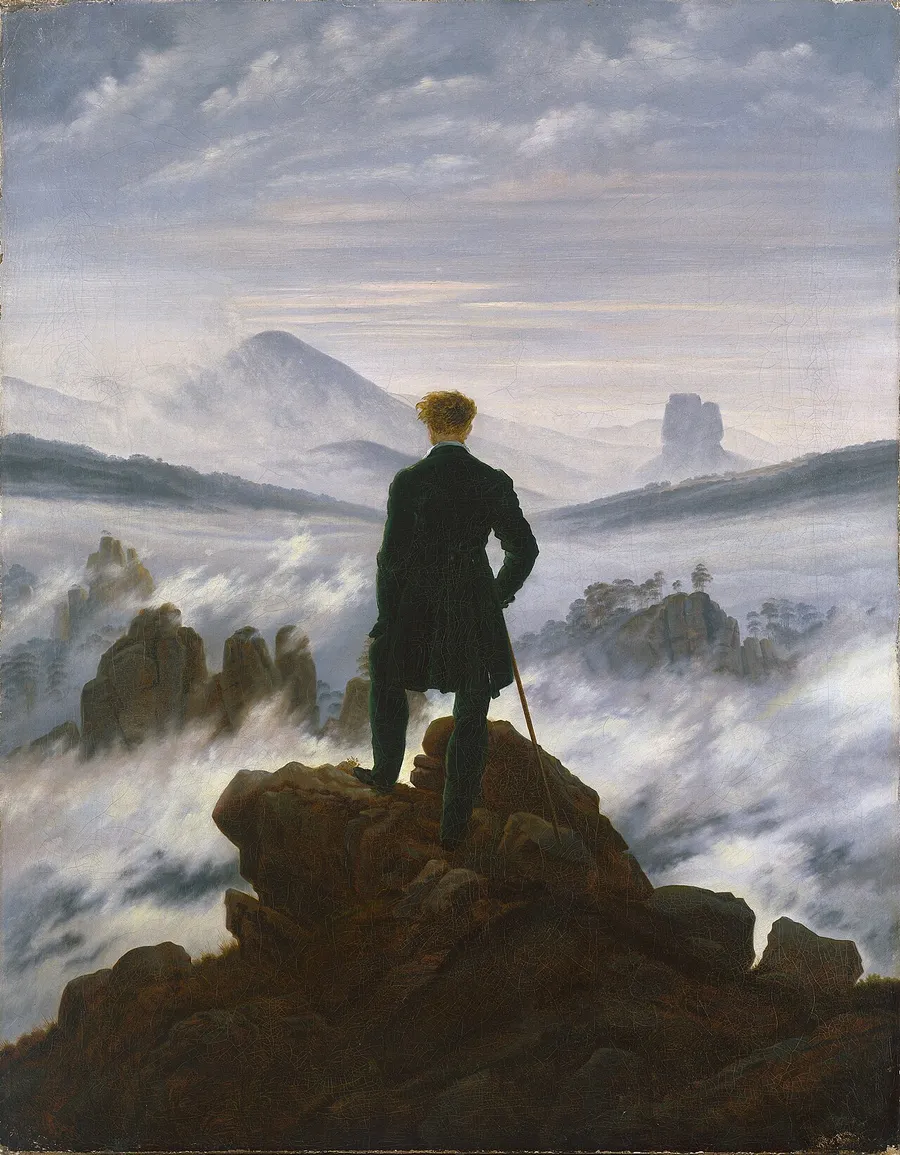 A man in a dark coat stands on a rocky summit with his back to us, looking out over a sea of fog with mountain peaks rising through it.